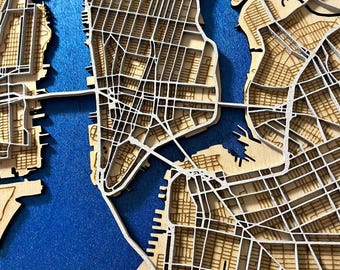 May include: A detailed, layered wooden map of New York City, featuring the city's grid layout and waterways. The map is made of light-colored wood with white lines representing streets, set against a blue background. Bridges and the city's outline are visible.