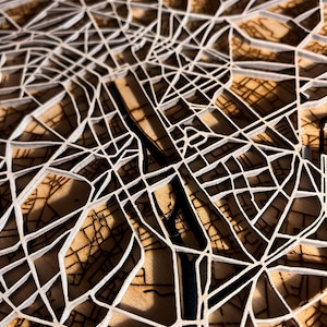 Könnte beinhalten: Eine detaillierte, mehrschichtige Holzkarte mit einem Netzwerk weißer Linien, die Straßen darstellen. Die Karte hat eine hellbraune Basis mit dunkleren braunen Details, wodurch ein 3D-Effekt entsteht. Das Design ist komplex und künstlerisch.