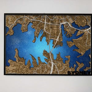 May include: A wooden map of Sydney, Australia, with a blue and gold color scheme. The map features a detailed depiction of the city's streets and waterways, with the word "SYDNEY" at the bottom. The map is framed in black.