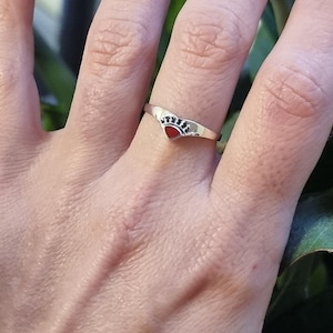 Puede incluir: Un anillo de plata con una pequeña piedra roja en forma de corazón engastada en el centro. El corazón está rodeado por una corona de plata.
