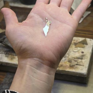 Puede incluir: Un pequeño colgante plateado brillante con la forma de un mapa de Palestina. El colgante se sostiene en la palma de una mano abierta. En primer plano, se ve una pulsera con un diseño en blanco y negro y un diseño de bandera. El fondo muestra una superficie de trabajo.