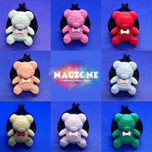 May include: A selection of teddy bear hair clips in various colours. Each clip features a bear with the word "Smile" and a bow tie. The bears are in shades of pink, red, green, and blue, set against a black background.