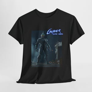 Puede incluir: Camiseta negra con un gráfico de un personaje de héroe oscuro de la serie Guyver. El personaje lleva un traje negro con un casco y está de pie en un entorno urbano oscuro. El texto "Guyver Dark Hero" está impreso en azul en la camiseta.