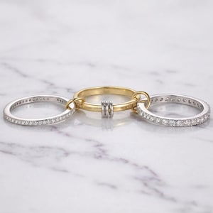 May include: Three connected rings. Two are silver, encrusted with small, clear stones. The central ring is gold, with two silver bands and a gold band. The rings are linked together, creating a unique jewelry design.
