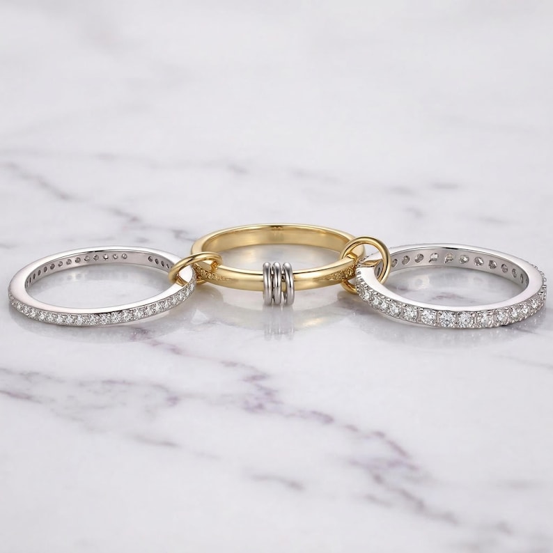 May include: Three connected rings. Two are silver, encrusted with small, clear stones. The central ring is gold, with two silver bands and a gold band. The rings are linked together, creating a unique jewelry design.