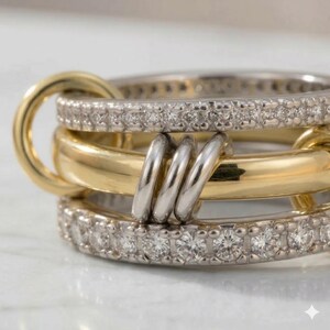 May include: Close-up of a multi-layered ring set. The ring features a gold band, a silver band with small, clear stones, and a silver band connecting the layers. The ring is on a white surface.