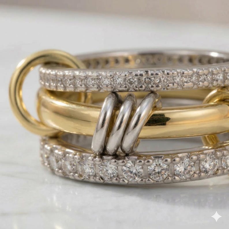 May include: Close-up of a multi-layered ring set. The ring features a gold band, a silver band with small, clear stones, and a silver band connecting the layers. The ring is on a white surface.