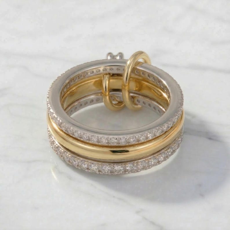 May include: A multi-band ring with a gold and silver design. The ring features three bands, with two silver bands encrusted with small, clear stones. The center band is gold. A gold loop connects the bands.