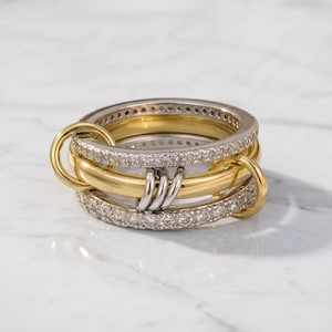 May include: A stack of gold and silver rings with a unique interlocking design. The rings feature a combination of smooth gold bands, silver bands with embedded clear stones, and connecting gold hoops. The rings are displayed on a white marble surface.