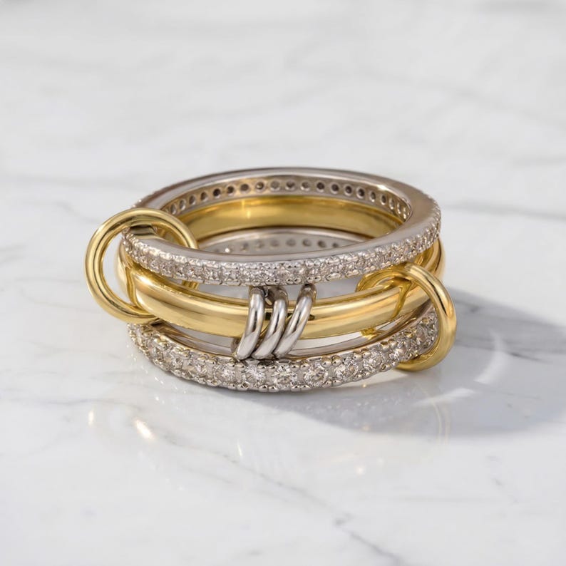 May include: A stack of gold and silver rings with a unique interlocking design. The rings feature a combination of smooth gold bands, silver bands with embedded clear stones, and connecting gold hoops. The rings are displayed on a white marble surface.