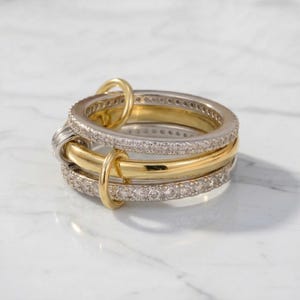 May include: A stack of three rings, two with a silver-tone finish and one with a gold-tone finish. The silver rings are encrusted with small, clear stones. The gold ring is smooth. The rings are interlocked with small gold connectors.