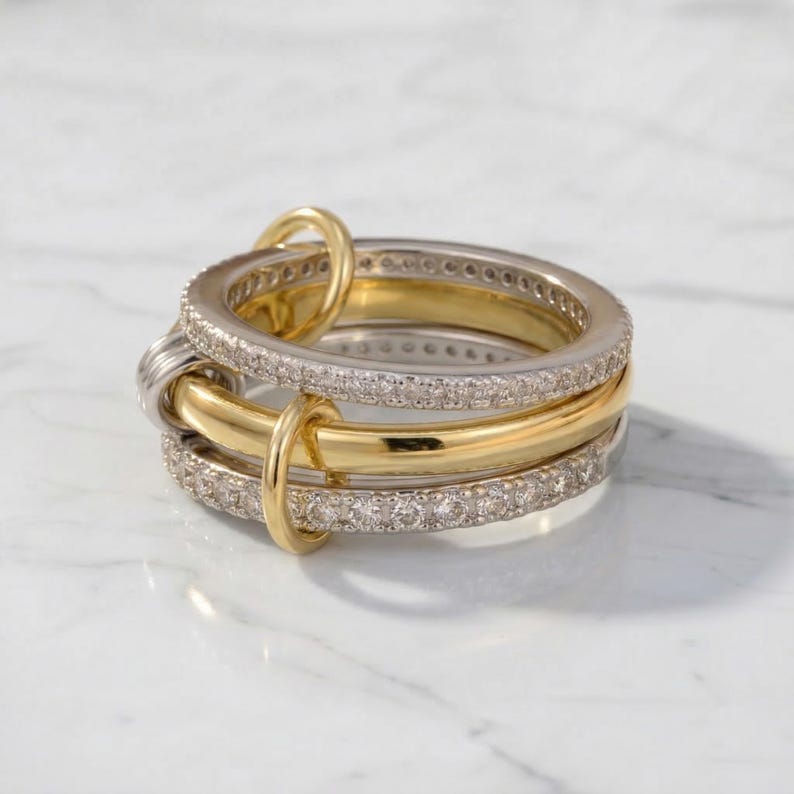 May include: A stack of three rings, two with a silver-tone finish and one with a gold-tone finish. The silver rings are encrusted with small, clear stones. The gold ring is smooth. The rings are interlocked with small gold connectors.