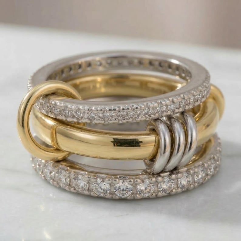 May include: A multi-band ring featuring a combination of gold and silver tones. The ring has three bands, with the top and bottom bands encrusted with small, clear stones. The middle band is gold, and the bands are interlocked.