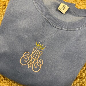 May include: A pale blue sweatshirt with a crew neck and long sleeves. The sweatshirt has an embroidered design of a crown above the intertwined letters "AM" in a light pink colour.
