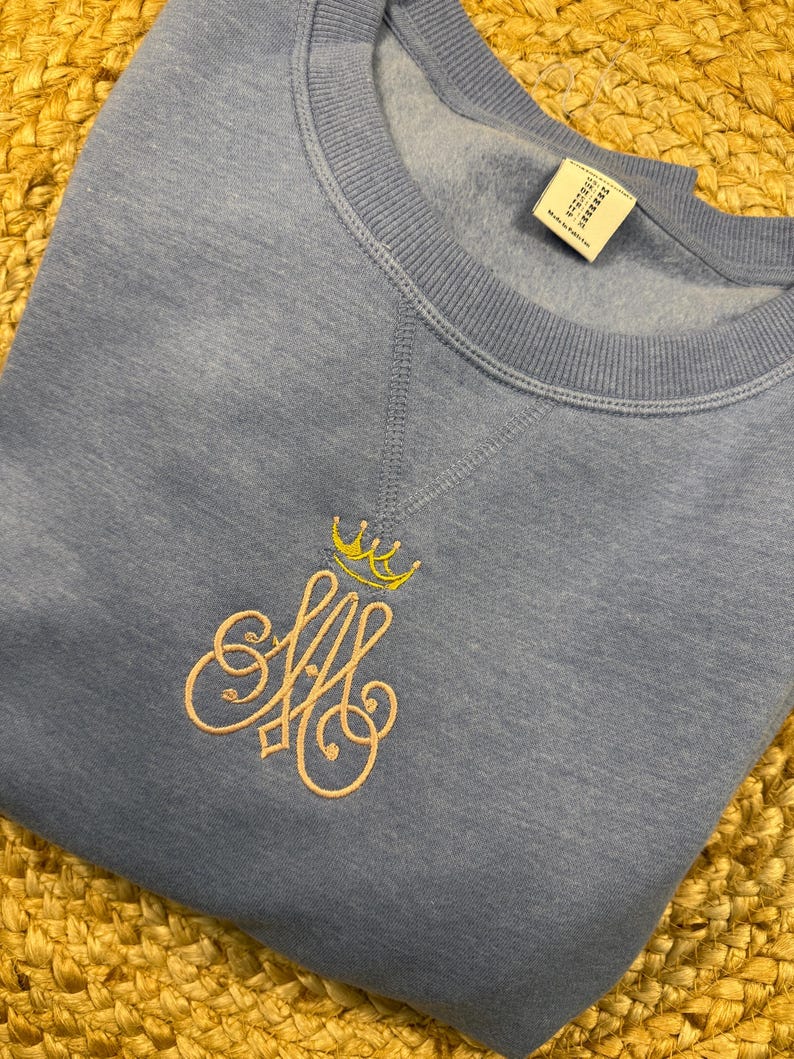 May include: A pale blue sweatshirt with a crew neck and long sleeves. The sweatshirt has an embroidered design of a crown above the intertwined letters "AM" in a light pink colour.