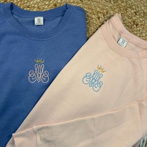 May include: Two crewneck sweatshirts, one light blue and one pink, are shown. Each has an embroidered monogram with a crown. The light blue sweatshirt has a gold crown and pink monogram. The pink sweatshirt has a gold crown and light blue monogram.