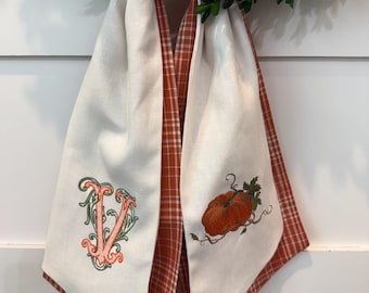 Può includere: Sciarpa in lino bianco con retro a quadri rossi e arancioni. Un lato presenta una zucca arancione ricamata, l'altro una lettera decorativa "V" in arancione e verde. La sciarpa è drappeggiata su una ghirlanda di foglie verdi.