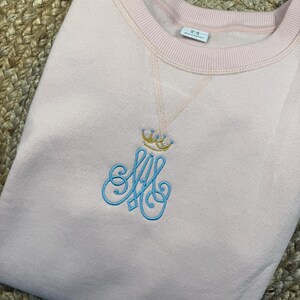 May include: A pale pink sweatshirt with a crew neck. The garment has an embroidered design of a crown above the intertwined letters "SA" in light blue. The sweatshirt is neatly folded on a woven surface.