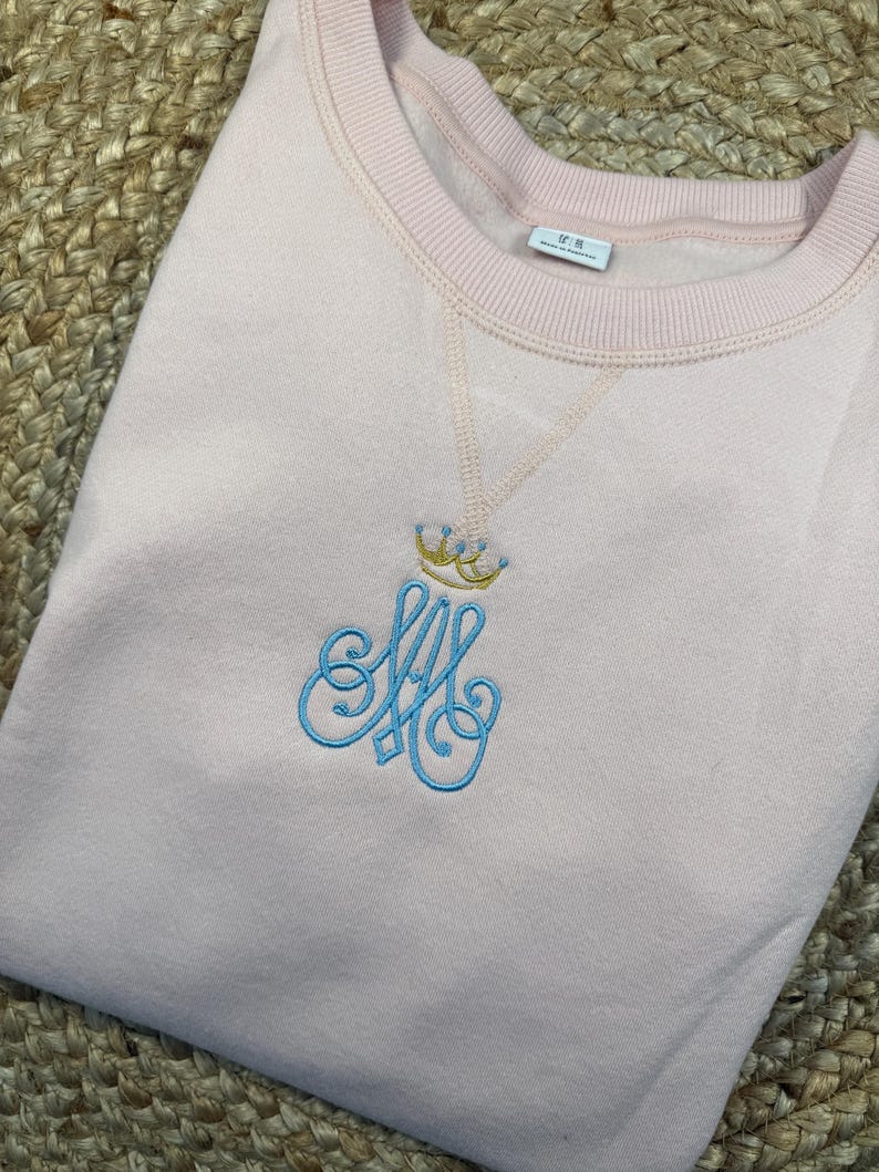 May include: A pale pink sweatshirt with a crew neck. The garment has an embroidered design of a crown above the intertwined letters "SA" in light blue. The sweatshirt is neatly folded on a woven surface.