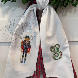 Puede incluir: Bufanda blanca con temática navideña con respaldo a cuadros rojos y verdes. Un lado presenta un cascanueces bordado, el otro un monograma verde y rojo. La bufanda está atada a una corona verde.