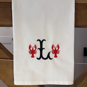May include: White hand towel with embroidered design. The design features a black letter "E" flanked by two red crayfish.