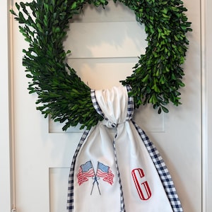 Könnte beinhalten: Ein grüner Buchskranz mit einem weiß-marineblauen Vichy-Band. Das Band ist mit zwei amerikanischen Flaggen und einem roten Buchstaben "G" bestickt. Dieser Kranz ist perfekt, um jedem Zuhause einen Hauch patriotischer Dekoration zu verleihen.
