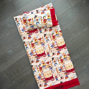 May include: A child's sleeping bag and pillow set with a cowboy theme. The set features a repeating pattern of cowboys, wagons, and musical instruments on a cream background. The edges are trimmed with red fabric, and the name "Gavin" is embroidered on the bottom.