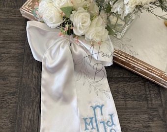 May include: A white satin ribbon bow featuring a cluster of white artificial roses and small pink accents. The long ribbon tails are embroidered with a light blue monogram featuring the letters M, G, and D in a decorative, swirling script font.
