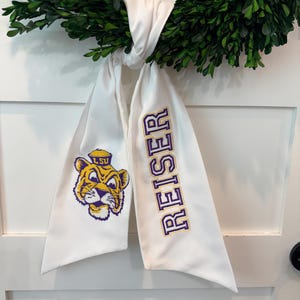 Può includere: Un nastro di tessuto bianco con la scritta "REISER" ricamata in viola e oro, e un logo di tigre dei cartoni animati con "LSU" su un cappello. Il nastro è legato a una ghirlanda verde, creando un accento decorativo.