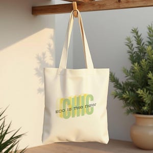 Peut inclure: Sac fourre-tout crème avec les mots "ECO IS THE new CHIC" en vert et jaune. Le sac est suspendu à un crochet en bois. Une plante en pot est visible en arrière-plan.