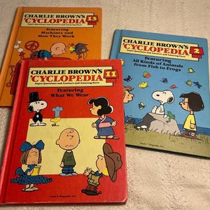 May include: Three hardback books titled Charlie Brown's Cyclopedia, volumes 2, 11, and 13. The orange volume 13 features machines, the red volume 11 shows various clothing, and the blue volume 2 depicts animals, all with Peanuts characters.
