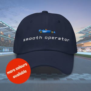 Może przedstawiać: Czapka bejsbolówka w kolorze granatowym z haftowanym białym samochodem wyścigowym i napisem "smooth operator". Czapka jest pokazana na rozmytym tle toru wyścigowego. Na pierwszym planie znajduje się czerwony okrąg z napisem "więcej kolorów dostępnych".