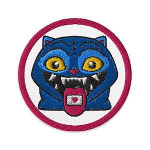 Könnte beinhalten: Gestickter Aufnäher mit einer blauen katzenartigen Kreatur mit großen gelben Augen, scharfen Zähnen und einer rosa Zunge mit einem herzförmigen Design. Der Aufnäher ist rund, mit einem weißen Zentrum und einem dunkelrosa Rand.
