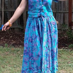 May include: A two-piece set consisting of a short-sleeved button-down top and a matching long, pleated skirt. Both pieces feature a vibrant turquoise blue base color with a repeating purple sun and celestial pattern throughout the fabric.