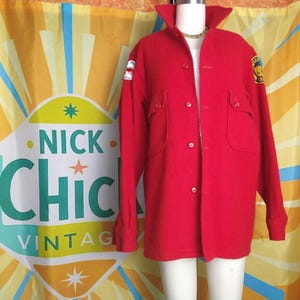 May include: A vintage red wool coat with a button-up front, two front pockets, and a wide collar. Patches are visible on the sleeves. The coat is displayed on a mannequin. The background features a colorful banner with the text "NICK CHIC VINTAGE."