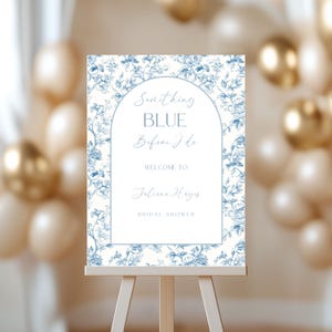 May include: A bridal shower welcome sign on a wooden easel. The sign features a floral design in blue and white, with the text "Something Blue Before I Do" and the name "Juliane Hayes".