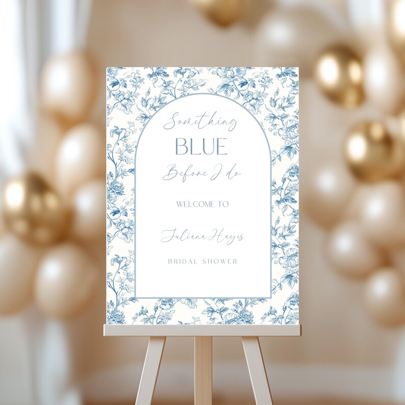 May include: A bridal shower welcome sign on a wooden easel. The sign features a floral design in blue and white, with the text "Something Blue Before I Do" and the name "Juliane Hayes".