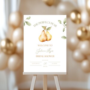 May include: A white sign on an easel with the text "THE PERFECT PEAR" and "WELCOME TO Juliana Hayes BRIDAL SHOWER". The sign features watercolor illustrations of pears and leaves, with event details.