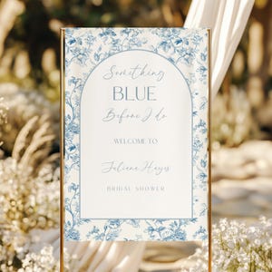 May include: A gold-framed sign with the text "Something Blue Before I Do" in elegant script. The sign features a floral design in shades of blue and white, with the name "Juliane Hayes" and "Bridal Shower" displayed below.