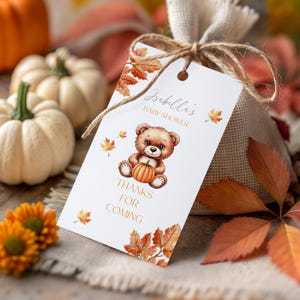 Op de afbeelding: Een bedankzakje voor een babyshower met een label met de tekst "Isabelli's Baby Shower" en "Thanks for Coming". Het label toont een teddybeer met een pompoen en herfstbladeren. De achtergrond bevat kleine pompoenen en herfstbladeren.