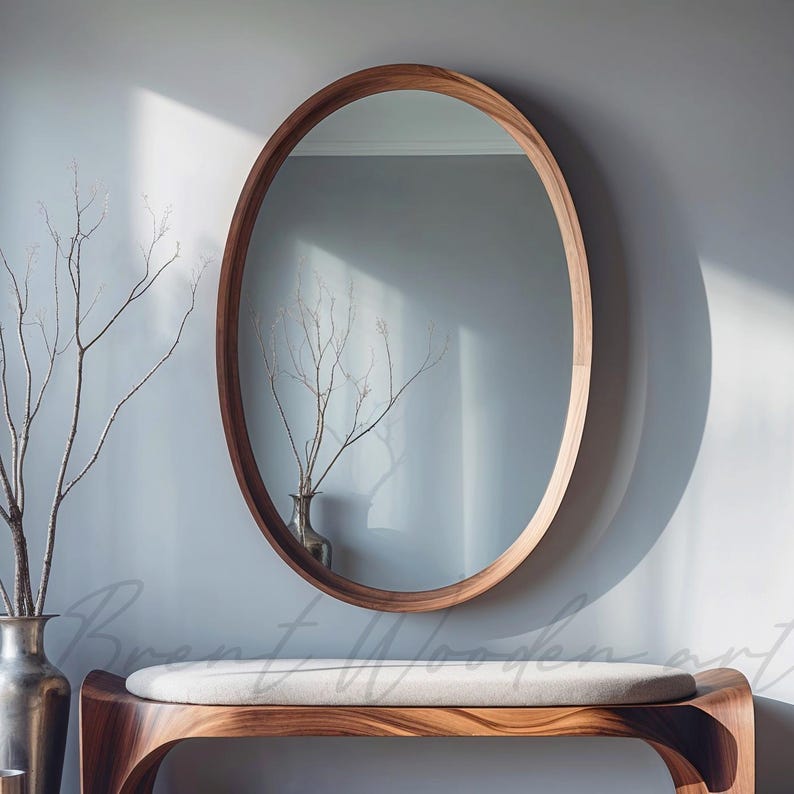 May include: A large oval mirror with a natural wood frame hangs on a light blue wall. The mirror reflects a room with a wooden bench and a vase of branches.