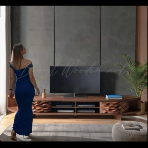 Pode incluir: Um móvel de TV moderno em madeira escura com uma grande televisão de tela plana. A unidade apresenta prateleiras abertas, frentes de gavetas decorativas em forma de onda e um perfil baixo. Uma mulher de vestido azul passa. O texto "Brent Wooden art" é visível.