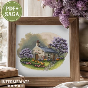May include: A framed cross-stitch of a cottage with a stone exterior, surrounded by purple flowering trees and colorful flowers. The artwork is in a wooden frame. Text on the image reads "PDF+ SAGA" and "INTERMEDIATE".