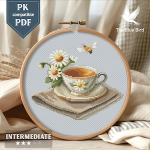 Op de afbeelding: Kruissteekborduurwerk van een kop en schotel met madeliefjes, een bij en een kopje op een servet. De borduursel zit in een houten hoepel. De tekst "PK compatible PDF" en "The Blue Bird" zijn zichtbaar.