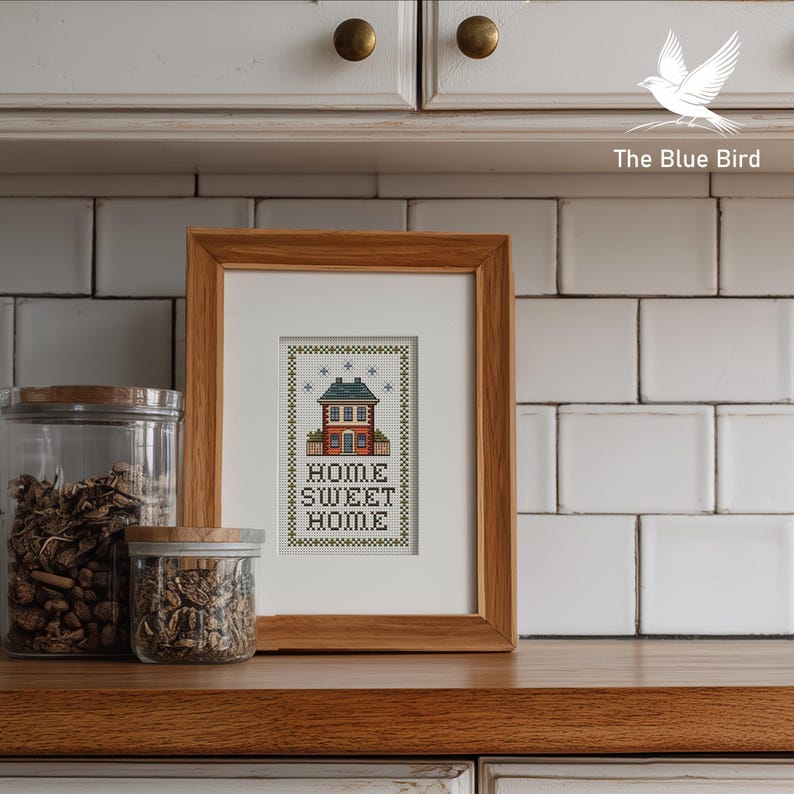 May include: A framed cross-stitch artwork with a wooden frame. The artwork depicts a house with the words "HOME SWEET HOME" below it. Two glass jars with wooden lids are on the left. The background is a white tiled wall.