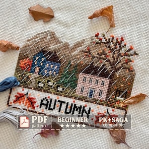 May include: A cross-stitch embroidery piece on white Aida cloth depicting an autumn landscape with a blue house, a pink house, trees, and the text AUTUMN. The design includes scattered dried brown leaves and loose blue and gray embroidery thread.