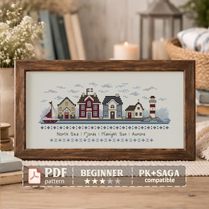May include: A framed cross-stitch pattern featuring a coastal village scene with houses, a sailboat, and a lighthouse. The artwork is in a wooden frame and includes the text "North Sea | Fjords | Midnight Sun | Aurora". The pattern is labeled "PDF BEGINNER".