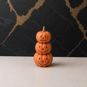 May include: A stack of three orange pumpkin-shaped candles, each featuring a unique carved jack-o'-lantern face with cut-out eyes, noses, and mouths. The top pumpkin has a small white wick protruding from its stem, and the entire structure is vertical.