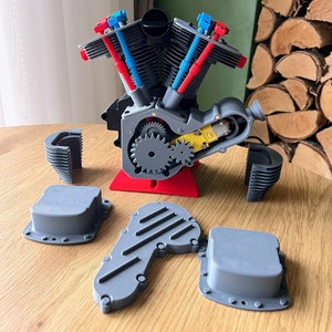May include: A 3D printed model of a V-twin engine with red, blue and grey parts. The engine is disassembled and the parts are laid out on a wooden surface.