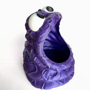 Może przedstawiać: Fioletowa doniczka w kształcie potwora z wypukłymi oczami i otwartą paszczą, wydrukowana w technologii 3D. Doniczka ma fakturowaną powierzchnię.